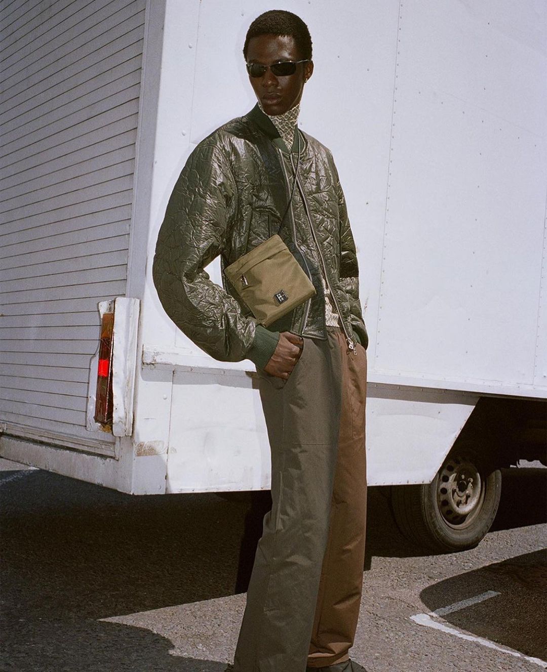An adult Black male poses outdoors wearing an olive quilted jacket, a crossbody bag, patterned turtleneck, and two-tone pants next to a white truck.