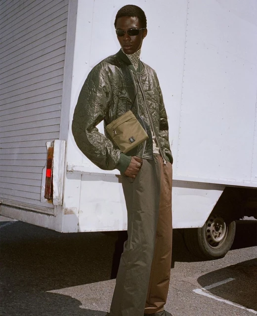 An adult Black male poses outdoors wearing an olive quilted jacket, a crossbody bag, patterned turtleneck, and two-tone pants next to a white truck.