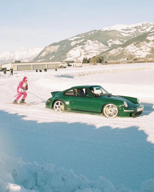A green Porsche is pulled by a person on skis across a snowy landscape, with a mountain range and clear blue sky in the background.