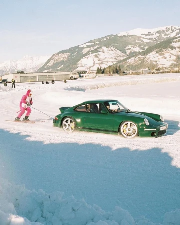 A green Porsche is pulled by a person on skis across a snowy landscape, with a mountain range and clear blue sky in the background.