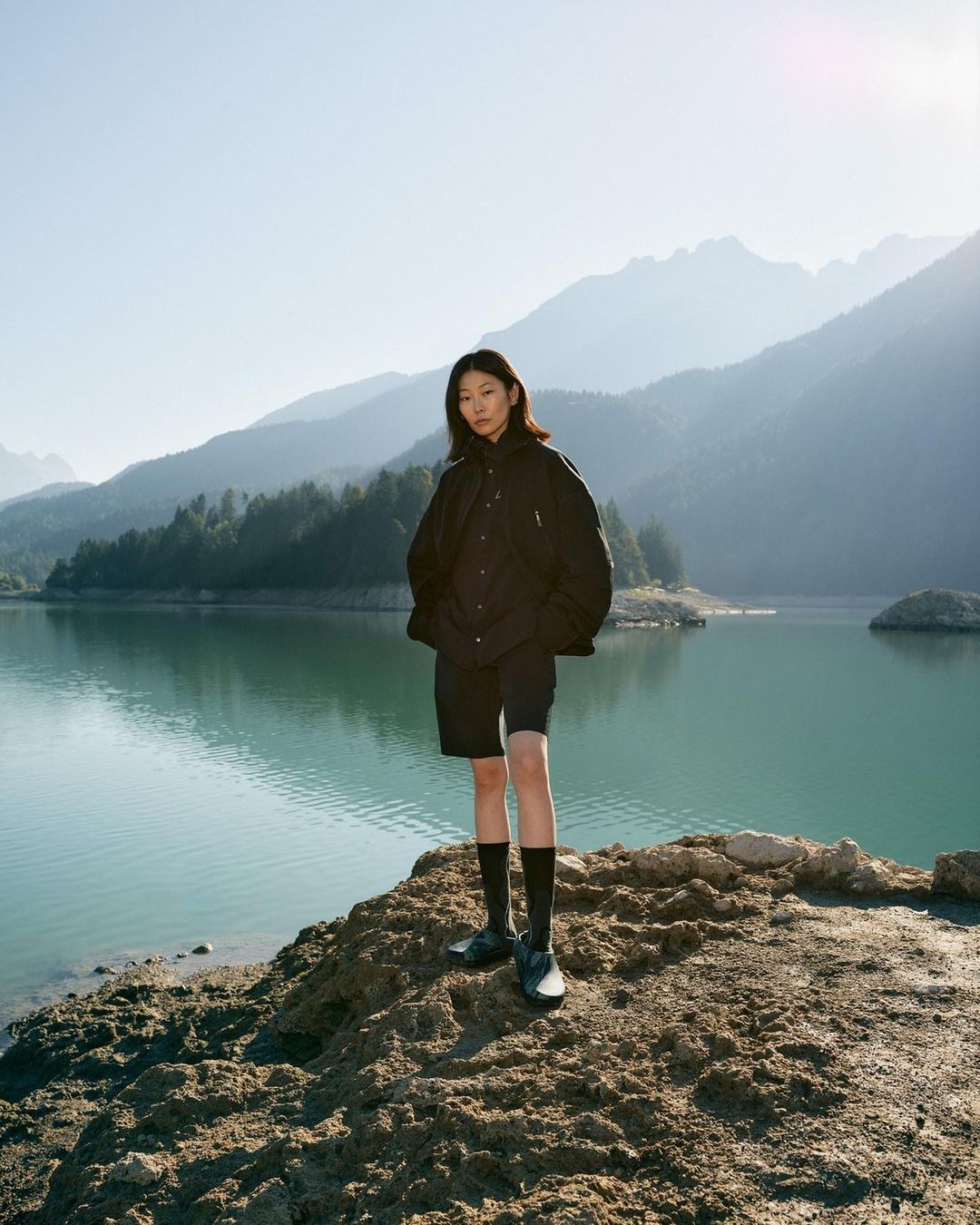 An Asian woman in a dark jacket and shorts poses on a rocky outcrop by a lake, with mountains and a clear blue sky in the background.