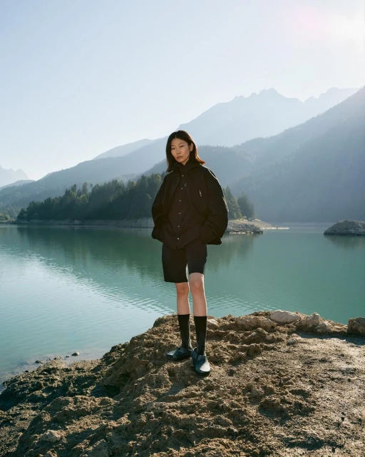 An Asian woman in a dark jacket and shorts poses on a rocky outcrop by a lake, with mountains and a clear blue sky in the background.