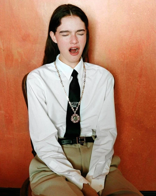 Woman in a white button-down shirt, black tie, and silver chain yawns with mouth open against a textured orange background.