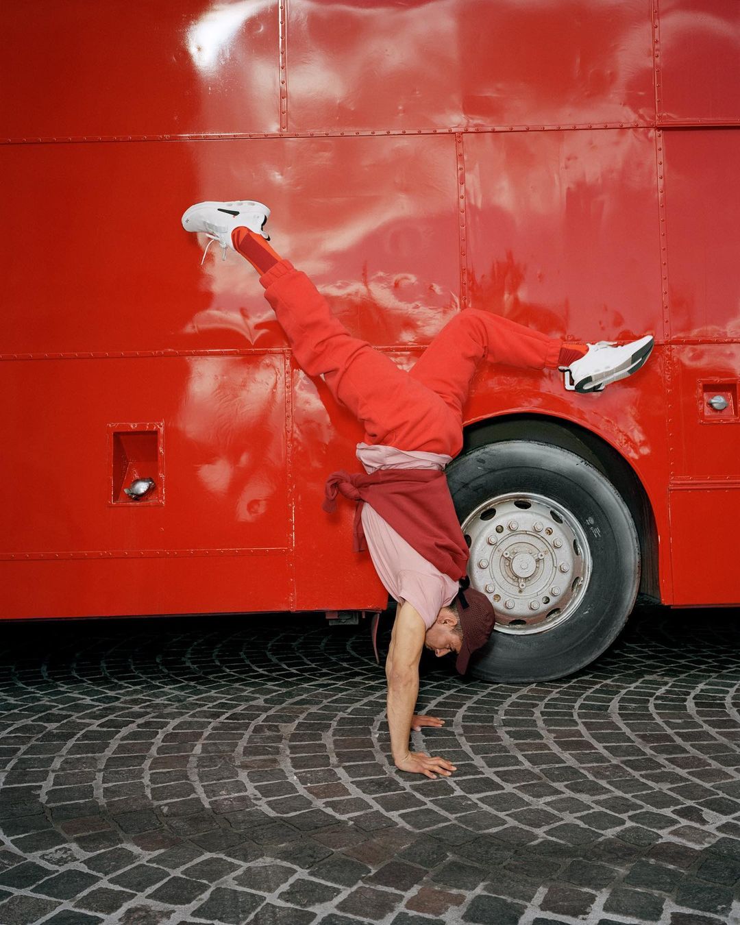 A person does a handstand in front of a red bus, wearing red pants and a hat, on a cobblestone street, showcasing fitness and urban style.
