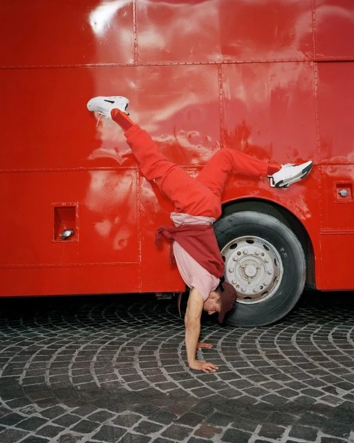 A person does a handstand in front of a red bus, wearing red pants and a hat, on a cobblestone street, showcasing fitness and urban style.