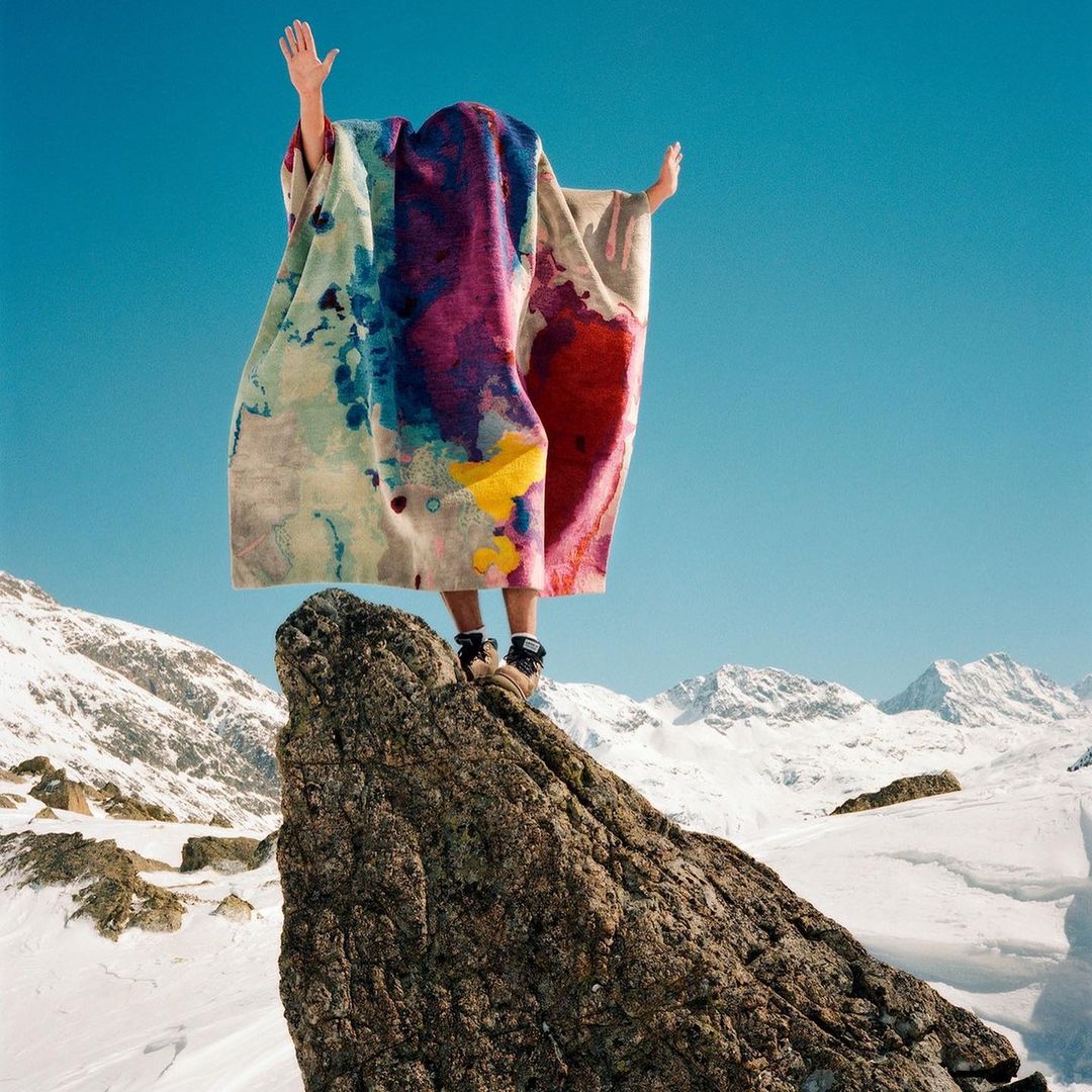 Person stands on snowy mountain peak beneath a colorful textile against blue sky, snow-covered peaks in the distance. Bright, outdoor setting.