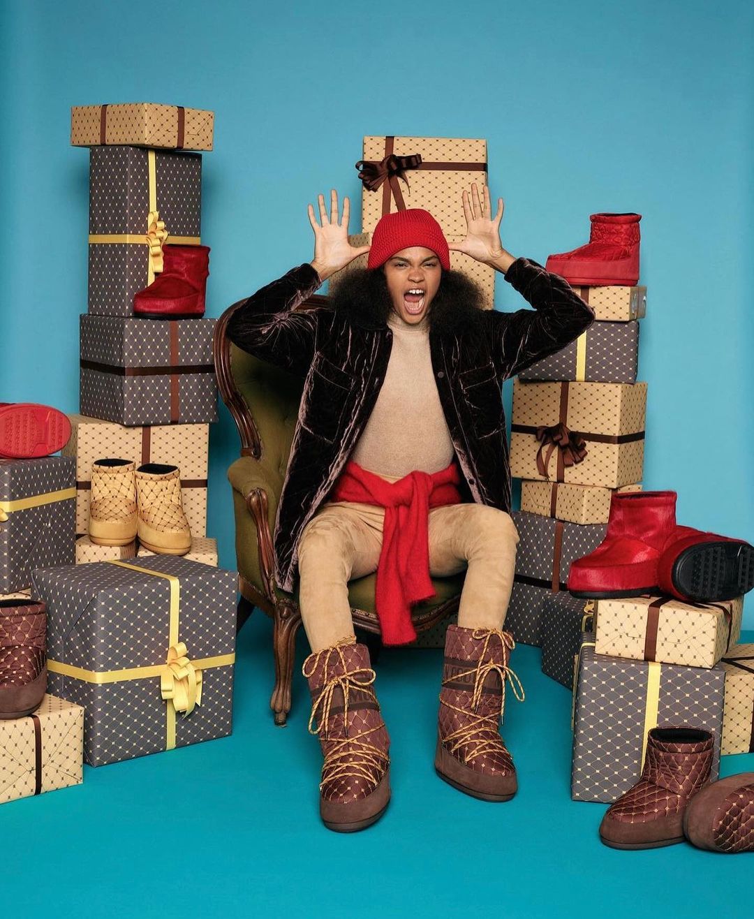 A young adult is sitting on a chair surrounded by many wrapped gifts and wearing winter boots, a red beanie, and a velvet jacket.