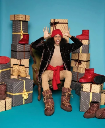 A young adult is sitting on a chair surrounded by many wrapped gifts and wearing winter boots, a red beanie, and a velvet jacket.