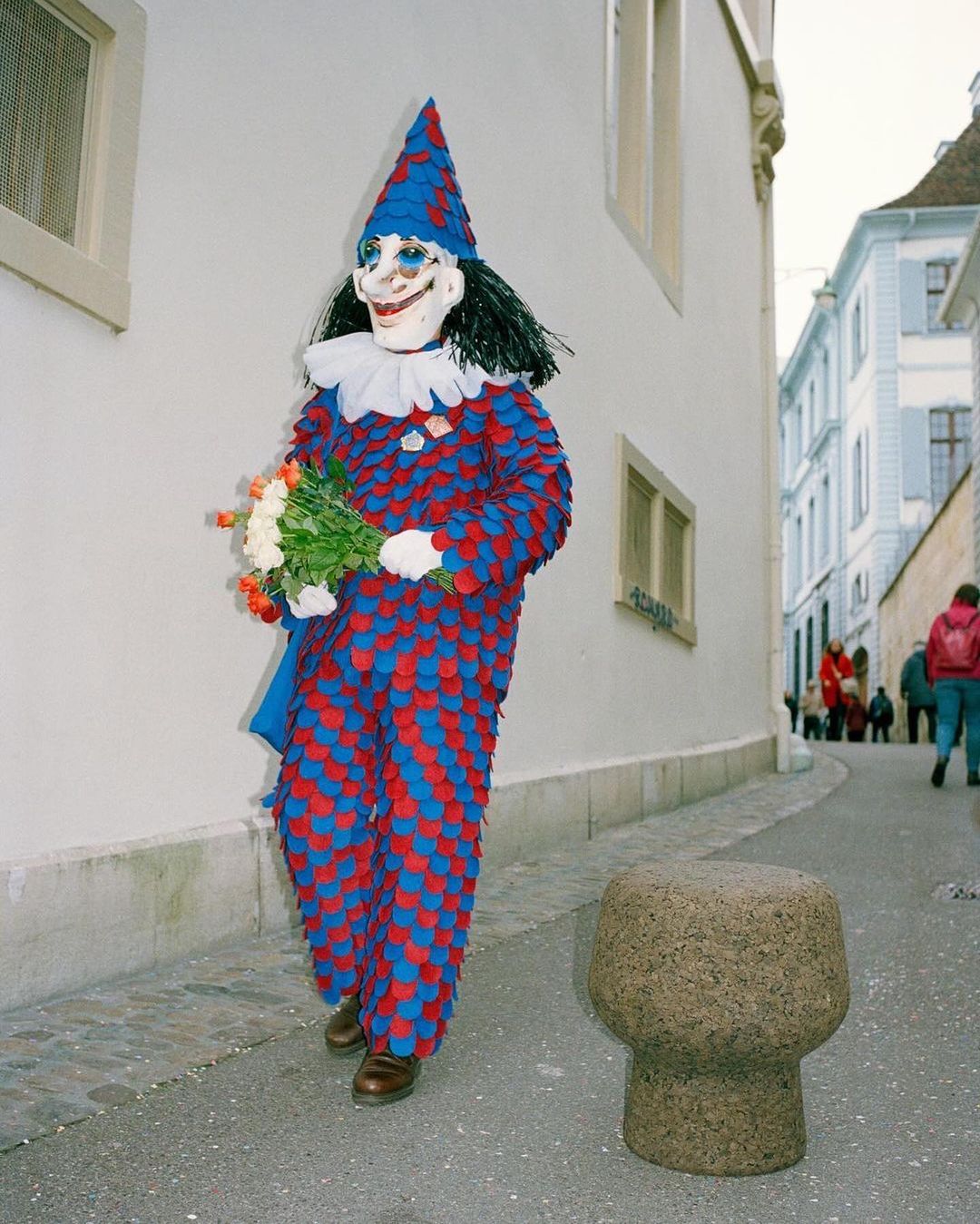 A person wearing a colourful clown costume and mask stands on a street holding a bouquet of flowers next to a stone bollard.