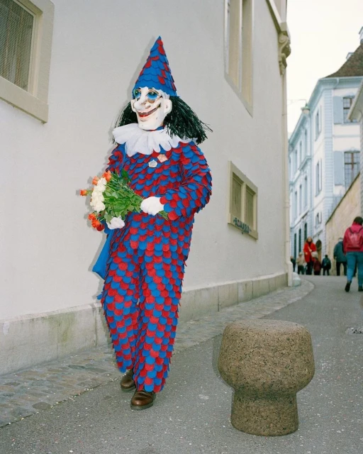 A person wearing a colourful clown costume and mask stands on a street holding a bouquet of flowers next to a stone bollard.