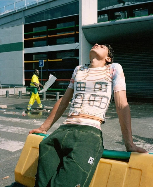 A young adult male relaxes on a yellow barrier outdoors on a sunny day, wearing a graphic tee and green pants, with a cityscape backdrop.