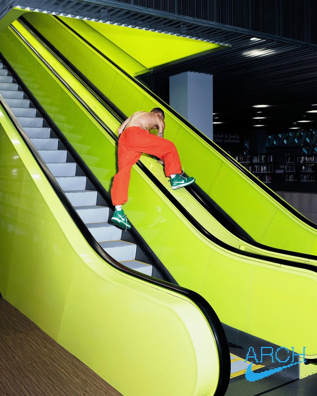 Athletic man climbing up the side of a modern yellow escalator, wearing red pants and green sneakers, inside a building with dark ceilings.