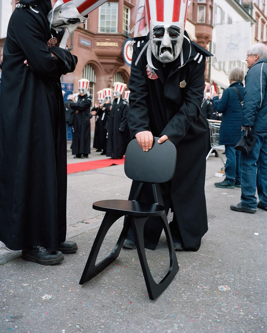 Group of people in costumes with masks and striped hats, one adjusting a black chair on a street, possible parade or festival.