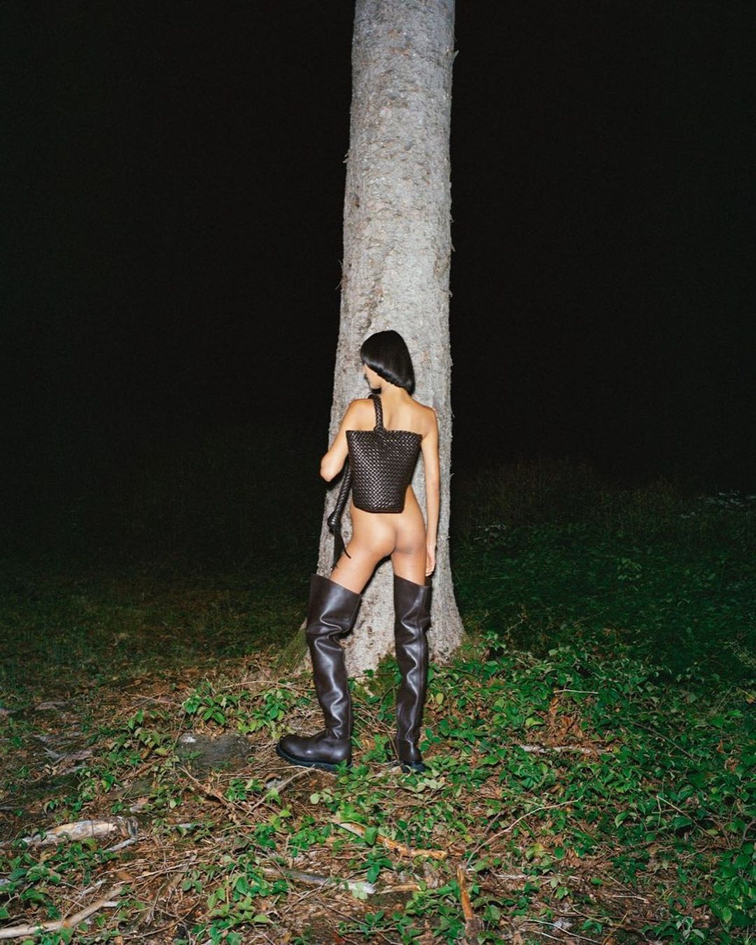 Back view of adult woman in boots posing near a tree in dark grassy environment with a woven bag and dark top.