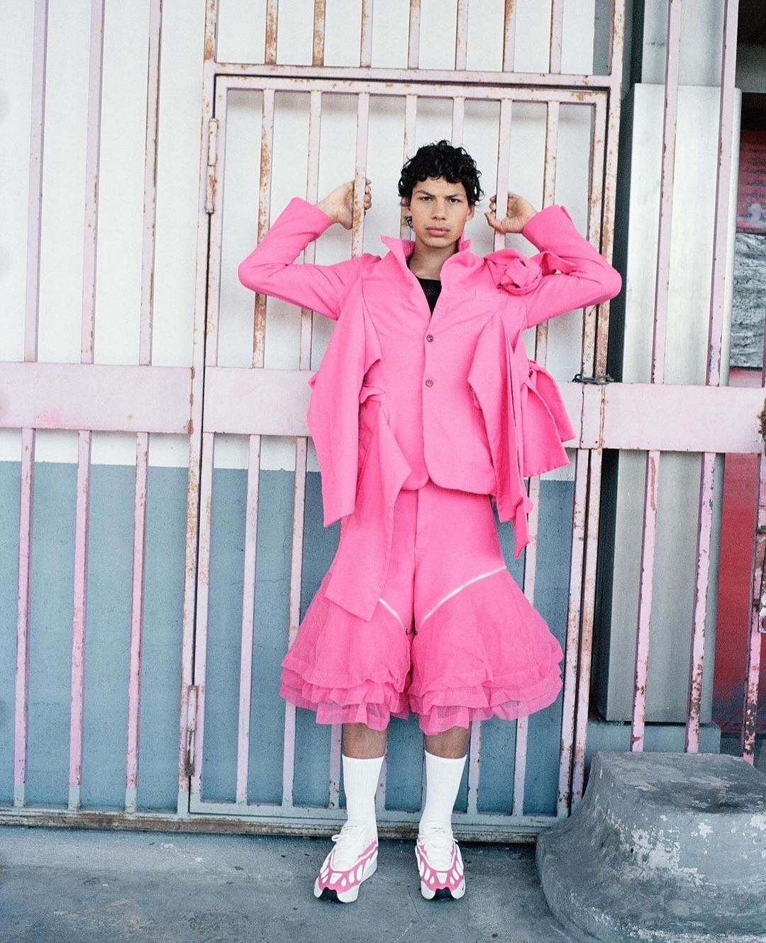 Young adult in pink suit poses near pink gate with white socks and modified roller skates, creating a bold fashion statement.