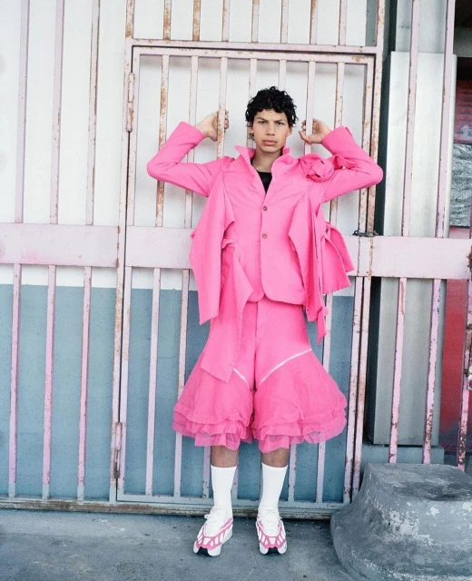 Young adult in pink suit poses near pink gate with white socks and modified roller skates, creating a bold fashion statement.