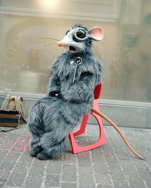 Person in a gray furry rat costume sitting on a red chair outdoors, holding a white stick. A tan accordion is visible nearby.
