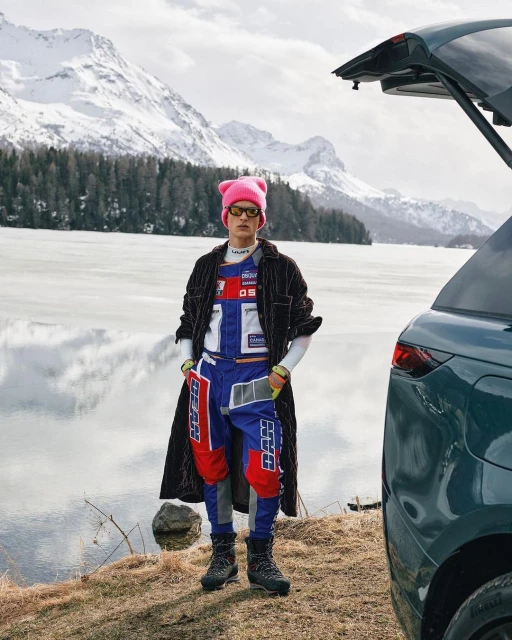 Fashionable adult male poses by a modern SUV with an open trunk, with snow-capped mountains and a frozen lake in the background, outdoor shot.
