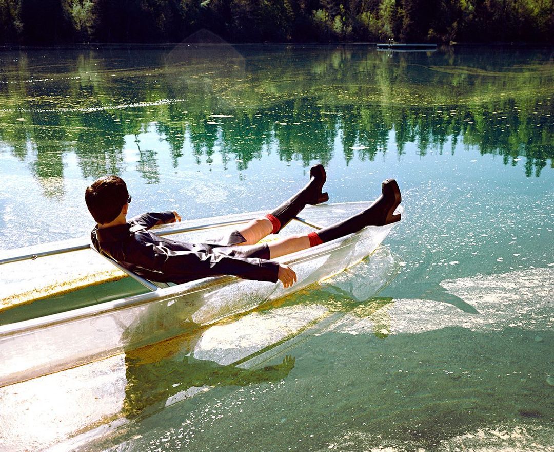 Person lies back in a transparent rowboat on a lake surrounded by green trees under a bright sky, captured in a serene outdoor setting.