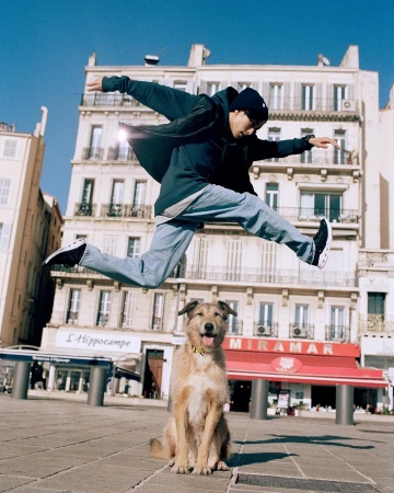 Adult male jumping in the air with a dog sitting on the ground; urban environment with building in background on a sunny day.
