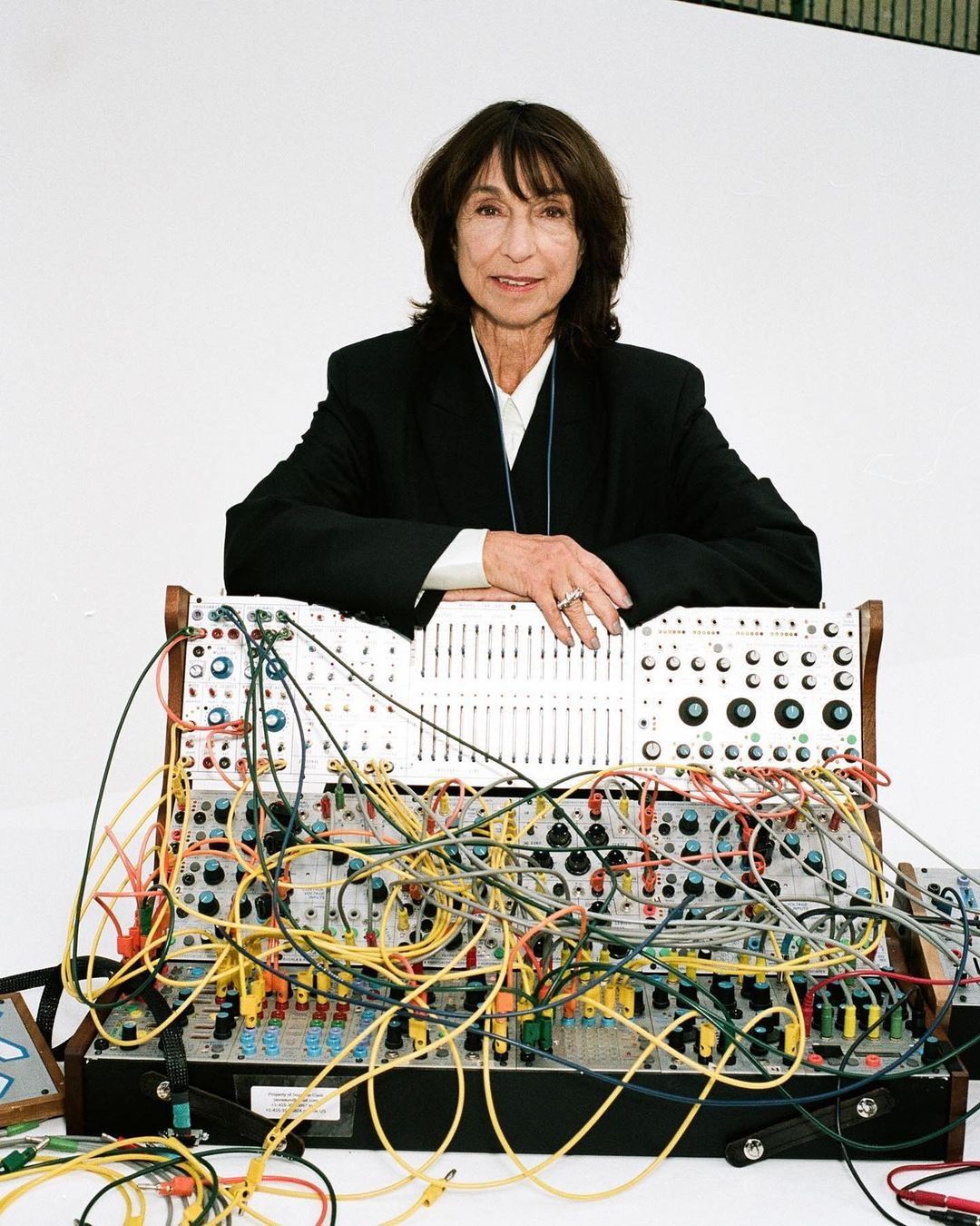 An older woman with short brown hair is standing behind a large modular synthesizer in a studio setting with a white background.