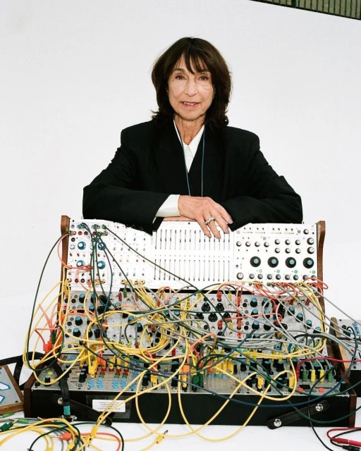 An older woman with short brown hair is standing behind a large modular synthesizer in a studio setting with a white background.