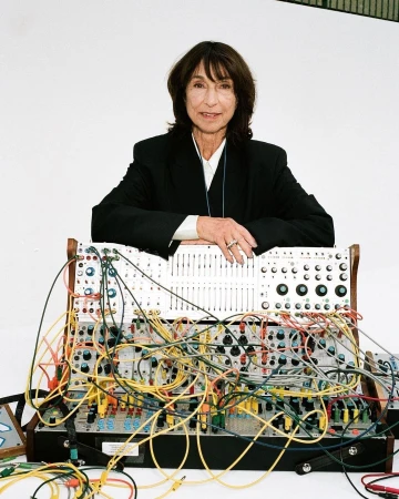 An older woman with short brown hair is standing behind a large modular synthesizer in a studio setting with a white background.