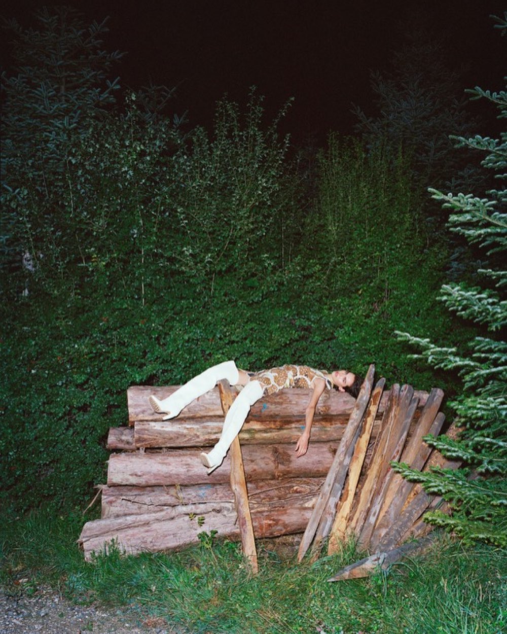 A person in white thigh-high boots and a sleeveless top reclines on logs in a dark forest setting at nighttime.
