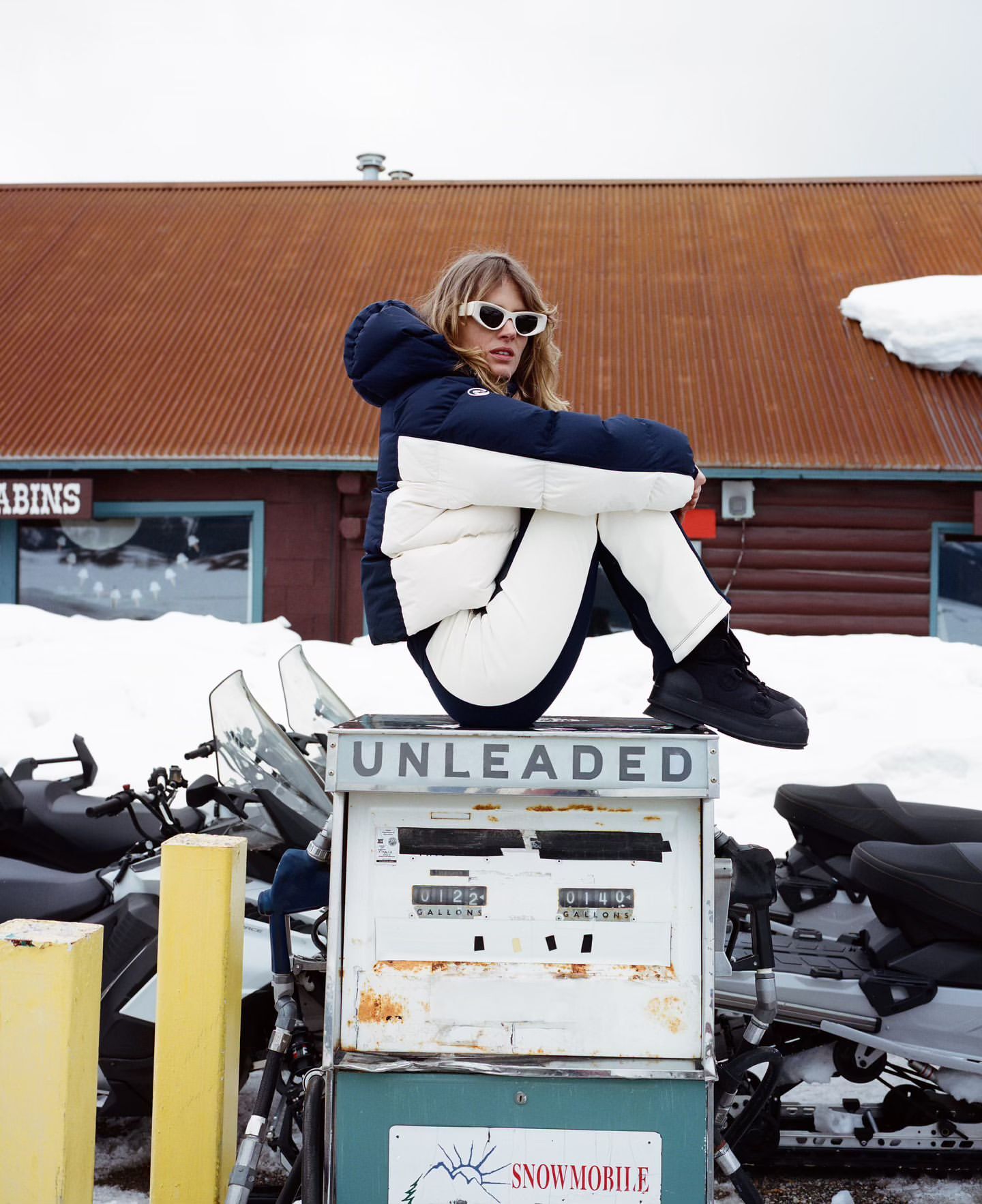 A blonde woman in winter clothing is seated atop an old gas pump with snowmobiles in the background, and a wooden building.