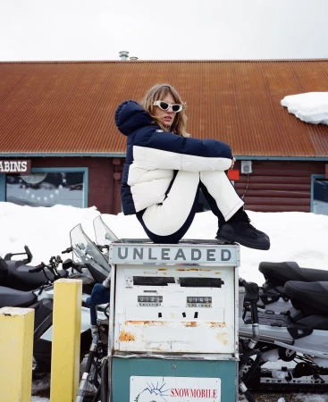 A blonde woman in winter clothing is seated atop an old gas pump with snowmobiles in the background, and a wooden building.