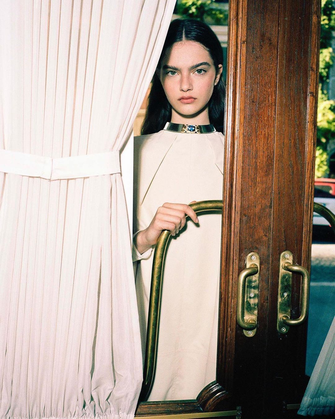Fashion shot of a brunette woman posing in a cream dress with a gold collar, holding a gold metal rail in a doorway next to white curtains.
