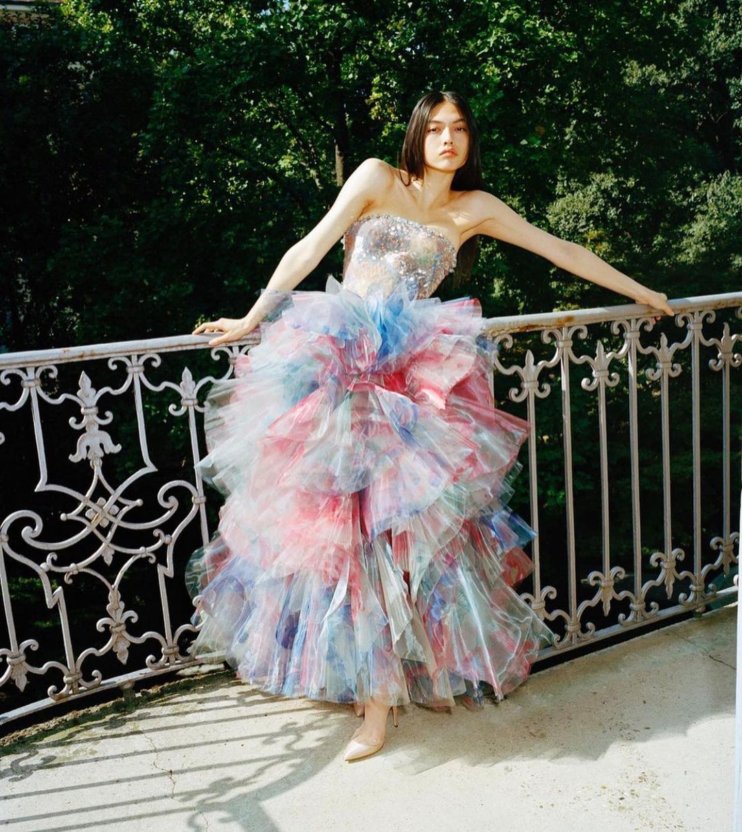 A young woman in a tiered, colorful tulle gown, poses on a balcony with ornate railings, with a verdant green backdrop of trees.