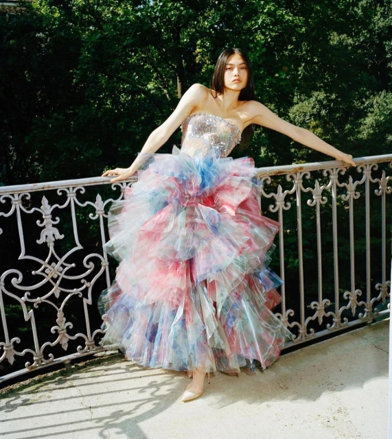 A young woman in a tiered, colorful tulle gown, poses on a balcony with ornate railings, with a verdant green backdrop of trees.