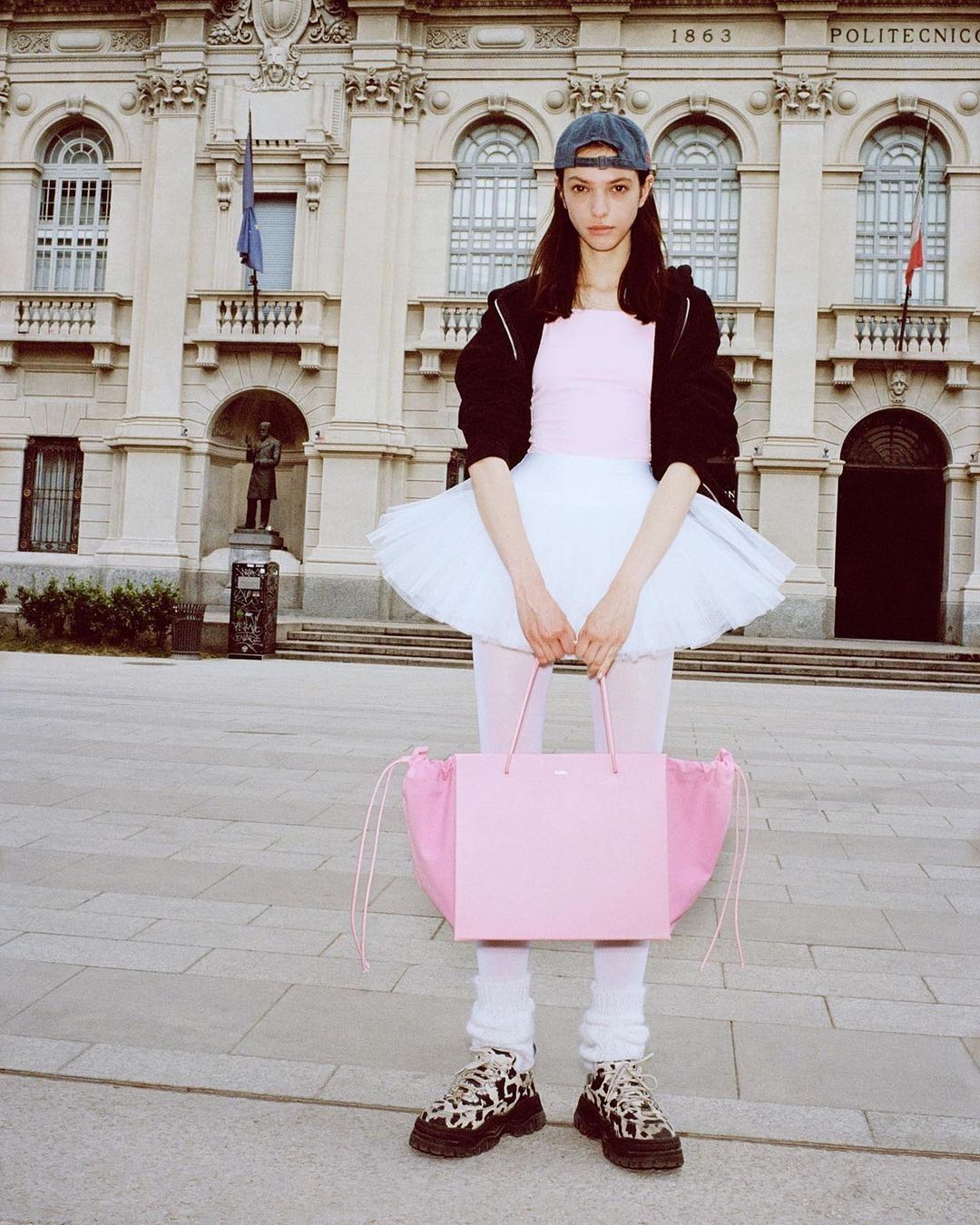 A woman in a tutu and baseball cap stands in front of an ornate building, holding a pink shopping bag, creating a surreal juxtaposition.