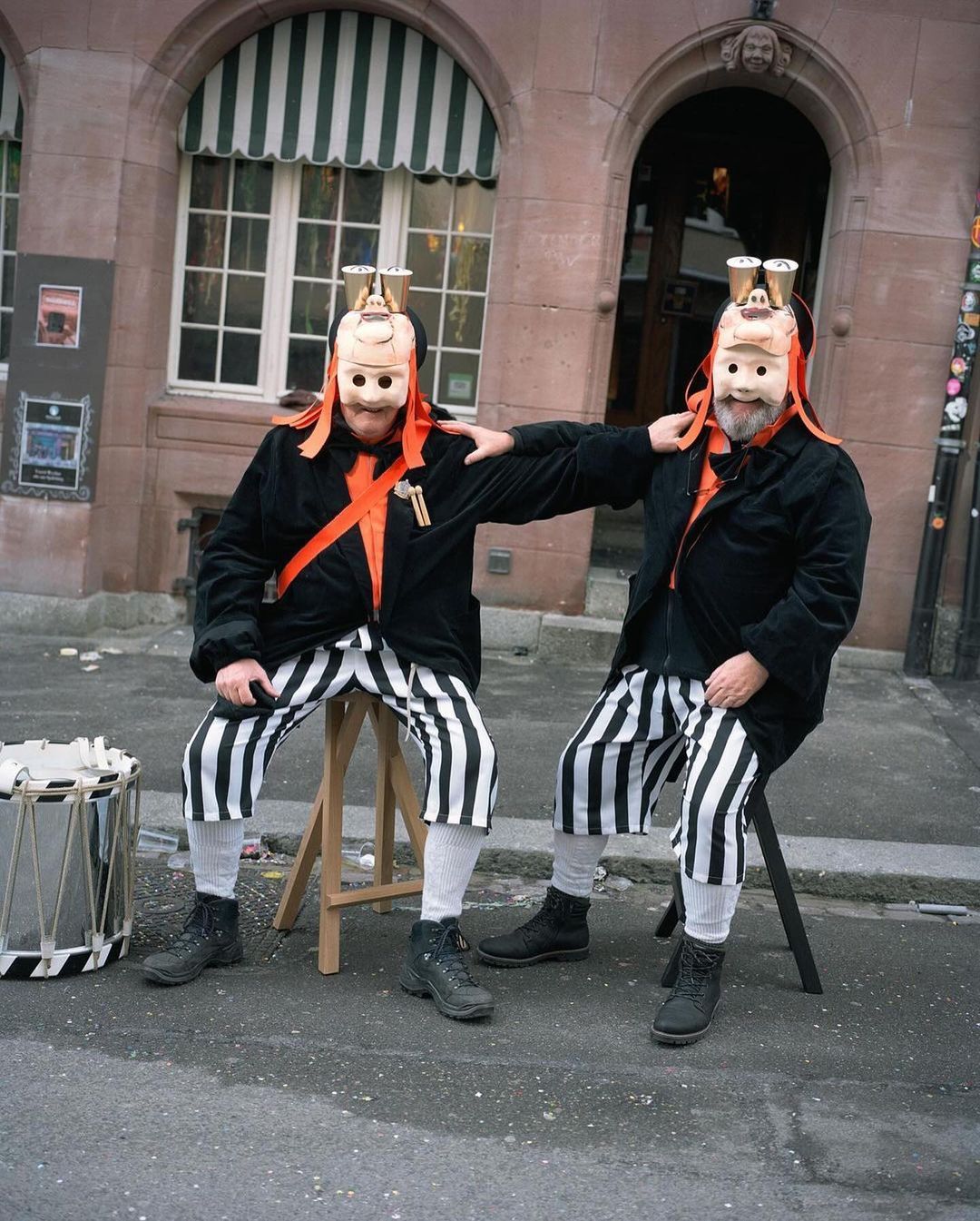 Two costumed men sit on stools outdoors. They wear black jackets, striped pants, orange sashes, and masks with metal attachments on their heads.