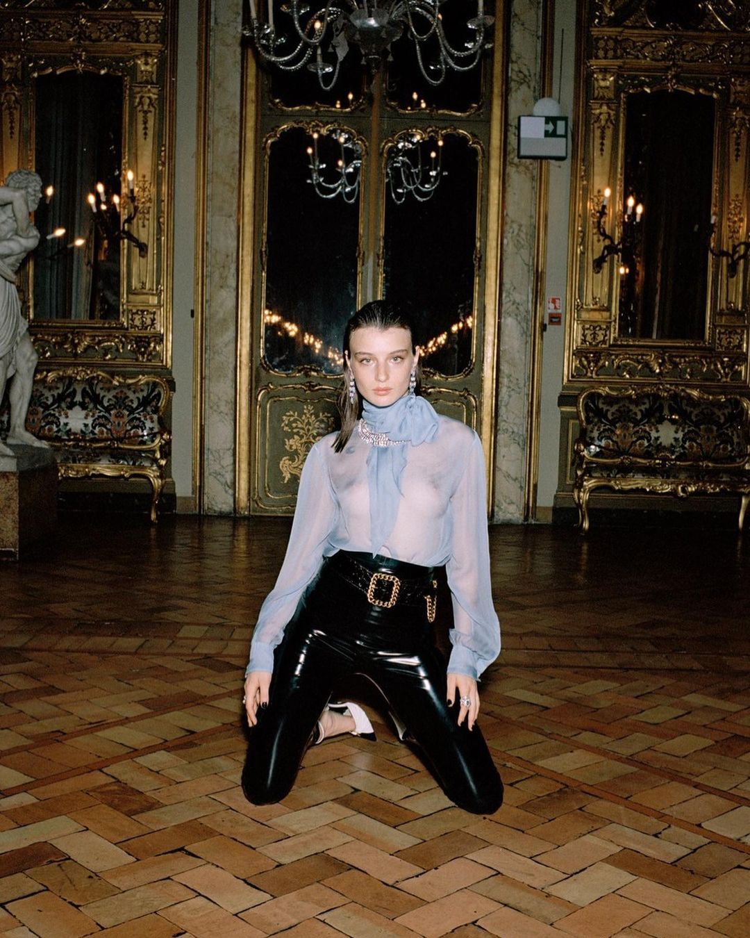 Elegant woman on knees in black patent leather pants and sheer blouse in ornate room with parquet floors and chandelier lighting.