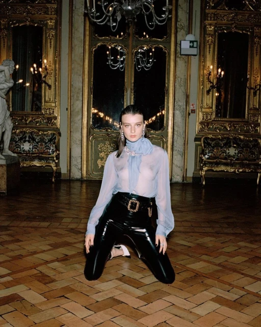 Elegant woman on knees in black patent leather pants and sheer blouse in ornate room with parquet floors and chandelier lighting.