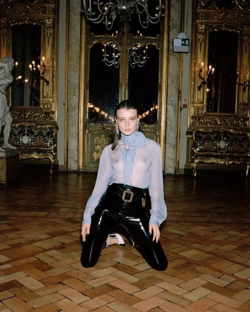 Elegant woman on knees in black patent leather pants and sheer blouse in ornate room with parquet floors and chandelier lighting.