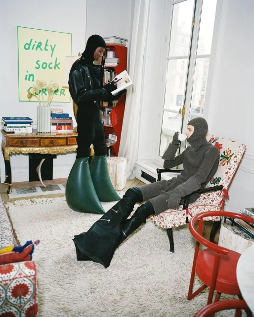 Two figures in full-body suits and oversized boots in an apartment, one seated holding a cup, the other standing reading a book.