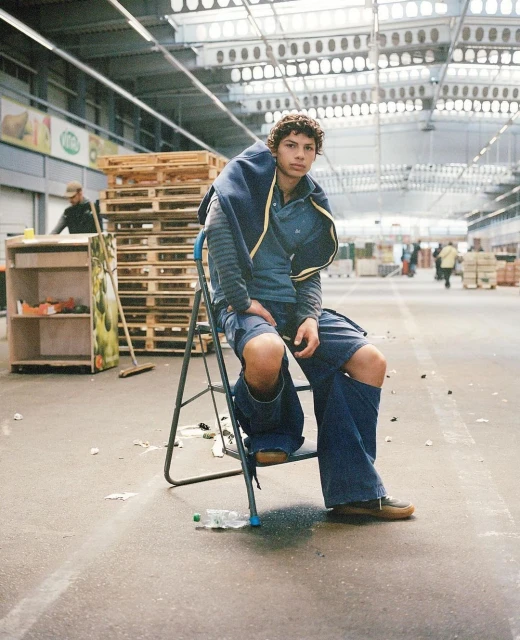 Young man in blue clothing sits on a stepladder in a warehouse with pallets and shelves in the background. The lighting is bright.