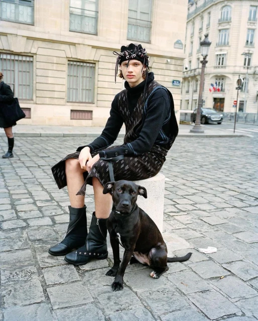 Young adult sits on a bollard in a city street wearing a black knit outfit with a headpiece, accompanied by a black dog.