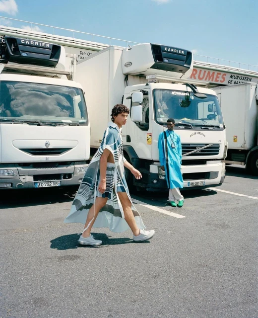 Two fashion models pose in front of white delivery trucks. One model walks in a matching cover-up and shorts, the other stands still.
