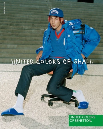 A man in a blue outfit and cap poses on a chair outside on a sunny day in an eye-level shot for a fashion advertising campaign.