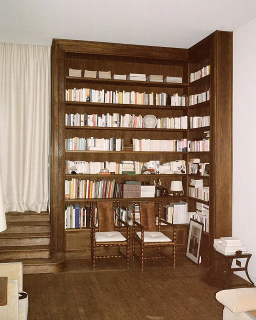 Interior view of a home library with floor-to-ceiling wooden bookshelves, two wicker chairs, a side table, and hardwood floors.