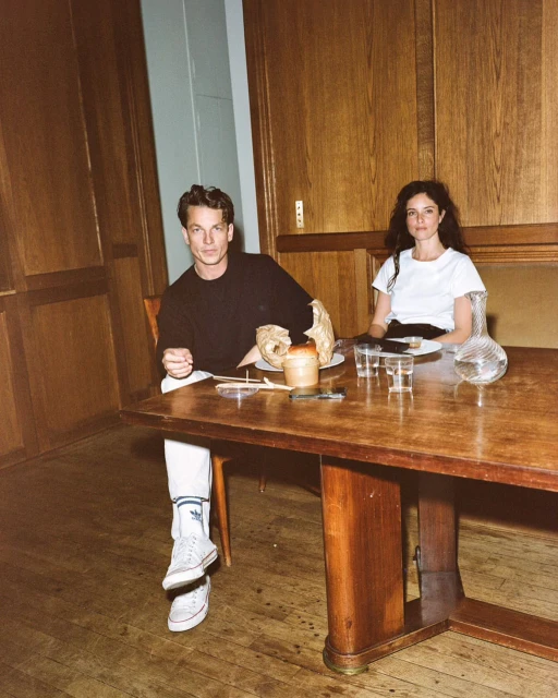 Indoor shot of a couple, an adult male and an adult female, sitting at a wooden table with dishware and a wooden paneled wall background.
