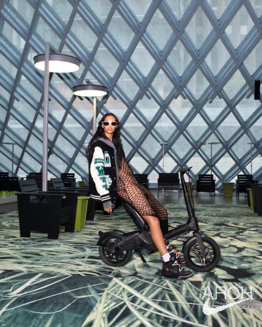 Woman in varsity jacket and fishnet stockings poses on black electric bicycle indoors against geometric glass wall backdrop.