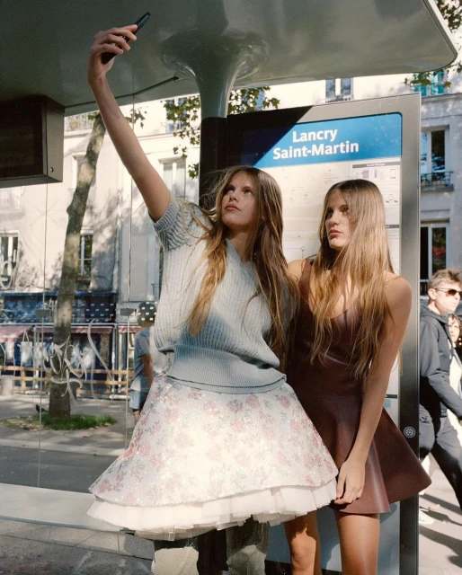 Two adult women pose for a selfie at a bus stop in an urban area; one is wearing a gray sweater and floral skirt, and the other a brown dress.