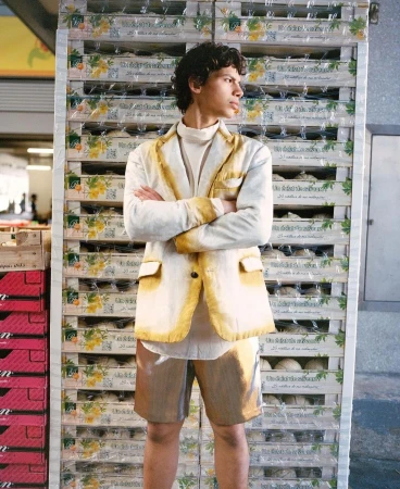 Young adult man poses with arms crossed in front of stack of boxes wearing a cream blazer, turtleneck, and metallic bronze shorts.