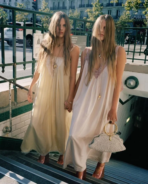 Two young women in long dresses hold hands on metro stairs in Paris, France. One carries a beaded bag; a city street is in the background.