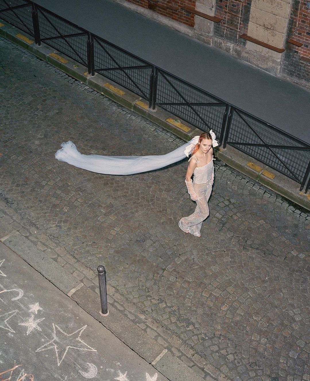 High-angle view of a woman wearing an embellished jumpsuit and veil on a cobblestone street in an urban setting at dusk.